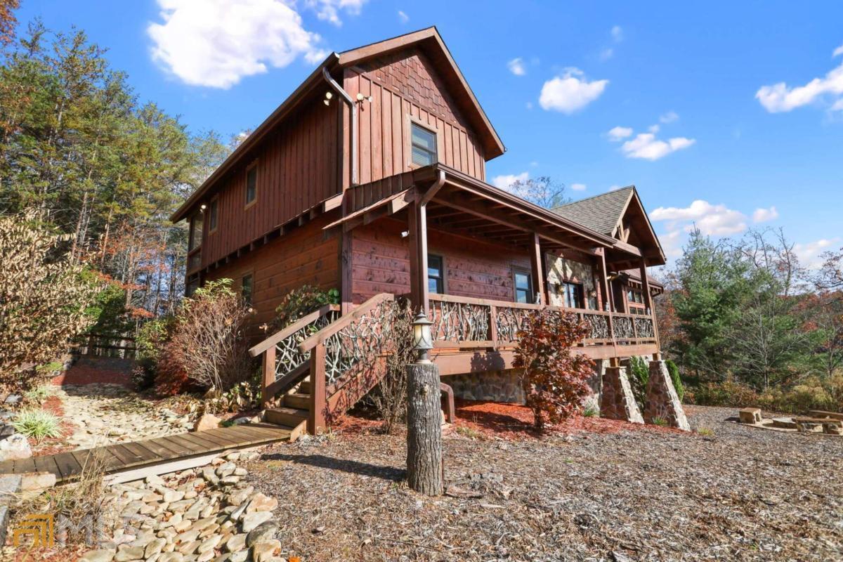 Rustic two-story wooden cabin with a wraparound porch and stone supports under a bright blue sky.