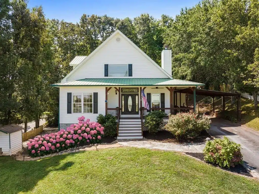White two-story house with a green metal roof, front porch, and pink hydrangea bushes in bloom.