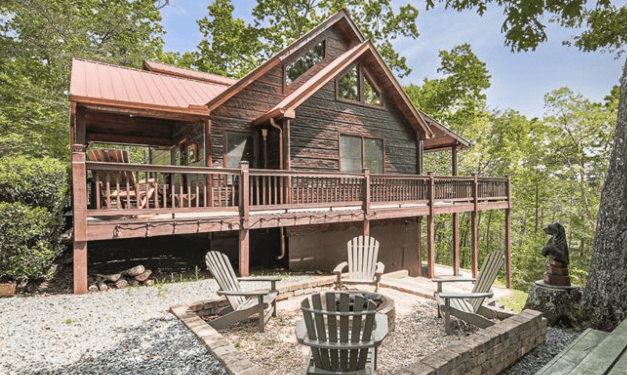 A wooden cabin on a raised deck in a forest, with Adirondack chairs around a fire pit.