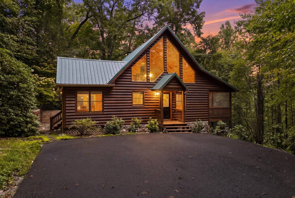 Cozy wooden log cabin with warm lights in a forest at sunset, driveway in foreground and trees surrounding the house
